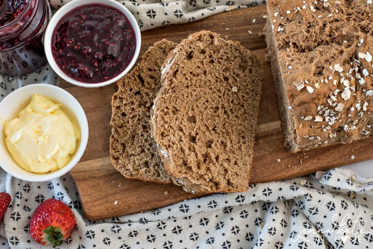 A few slices of brown bread resting on a wooden cutting board, next to small bowls of cream and jam