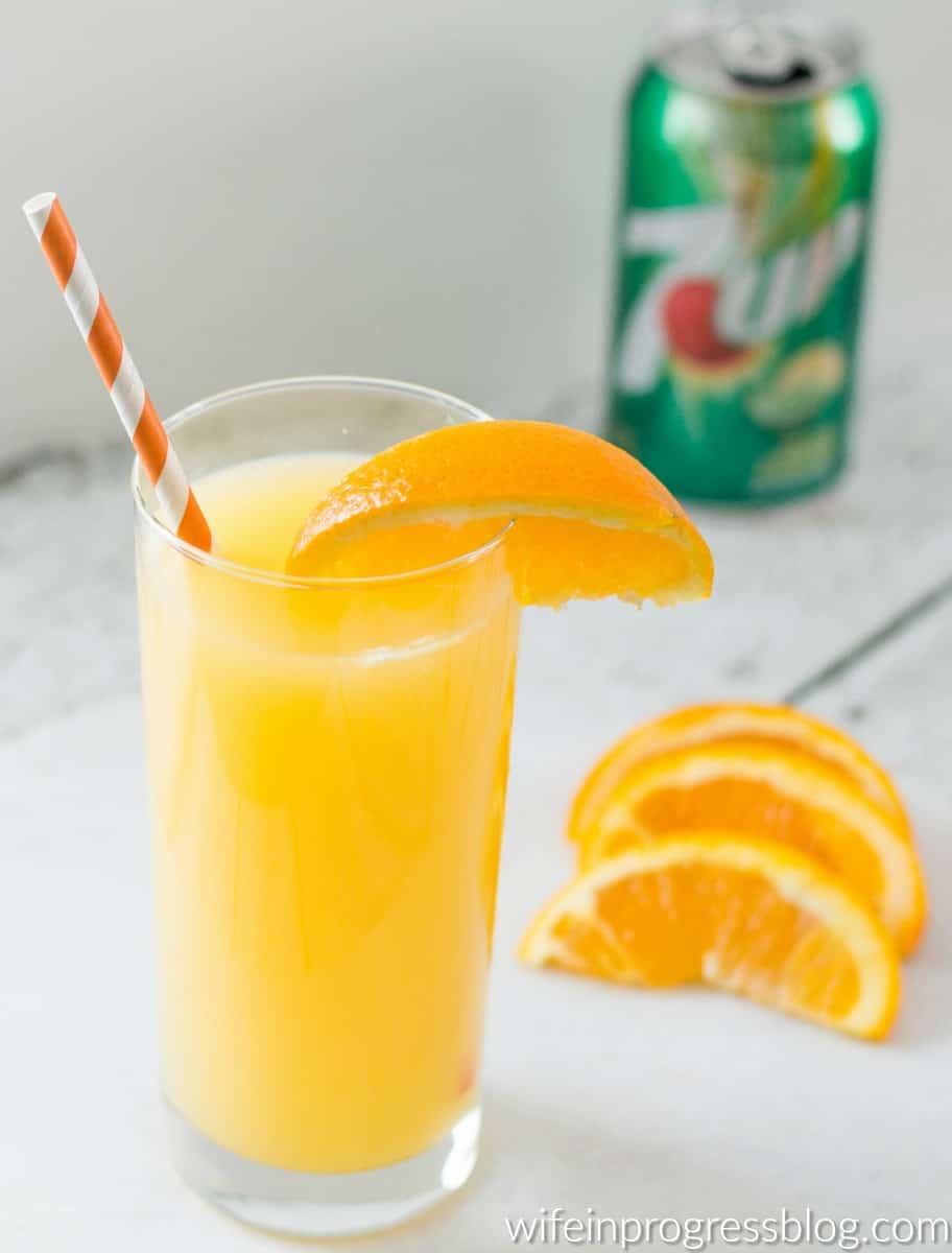 A glass of juice, with orange wedges and a can of 7UP soda nearby