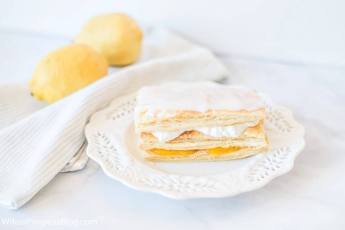 A lemon cream napoleon sitting on a dainty, white dessert plate with embossed edges, and a pair of lemons on a napkin nearby