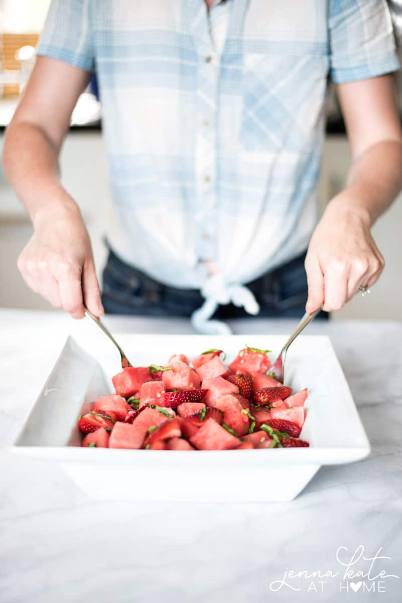 A person tossing a salad made of strawberry, watermelon and basil in a square, white bowl