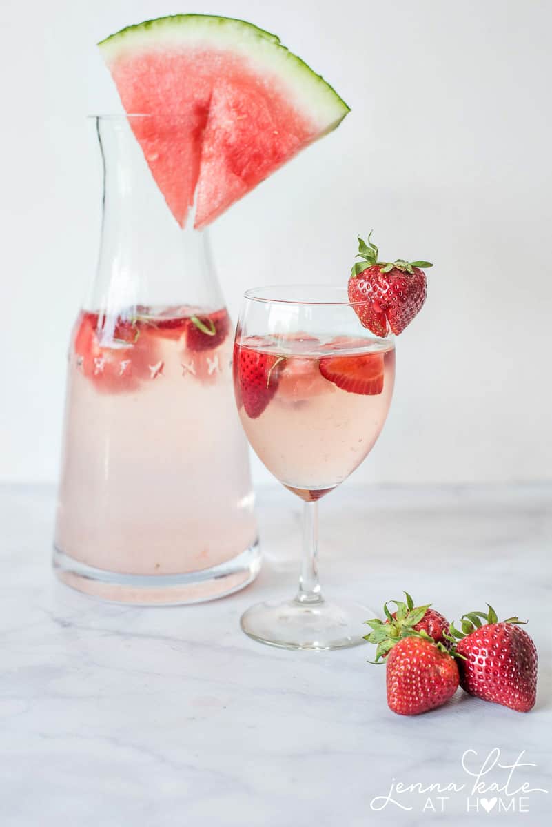 A clear glass pitcher and wine glass holding an alcoholic beverage, embellished with strawberries