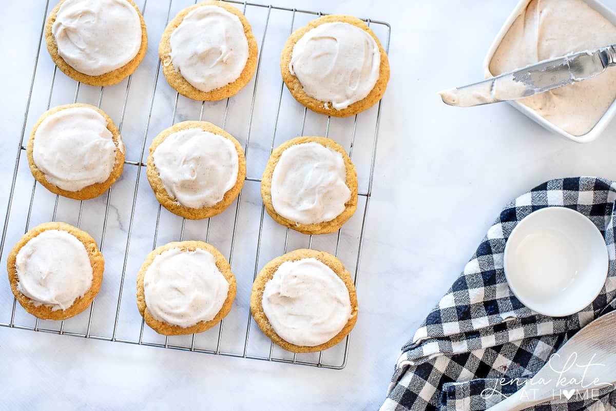 Pumpkin sugar cookies resting on cooling rack, with a bowl of cream cheese frosting and spreading knife nearby