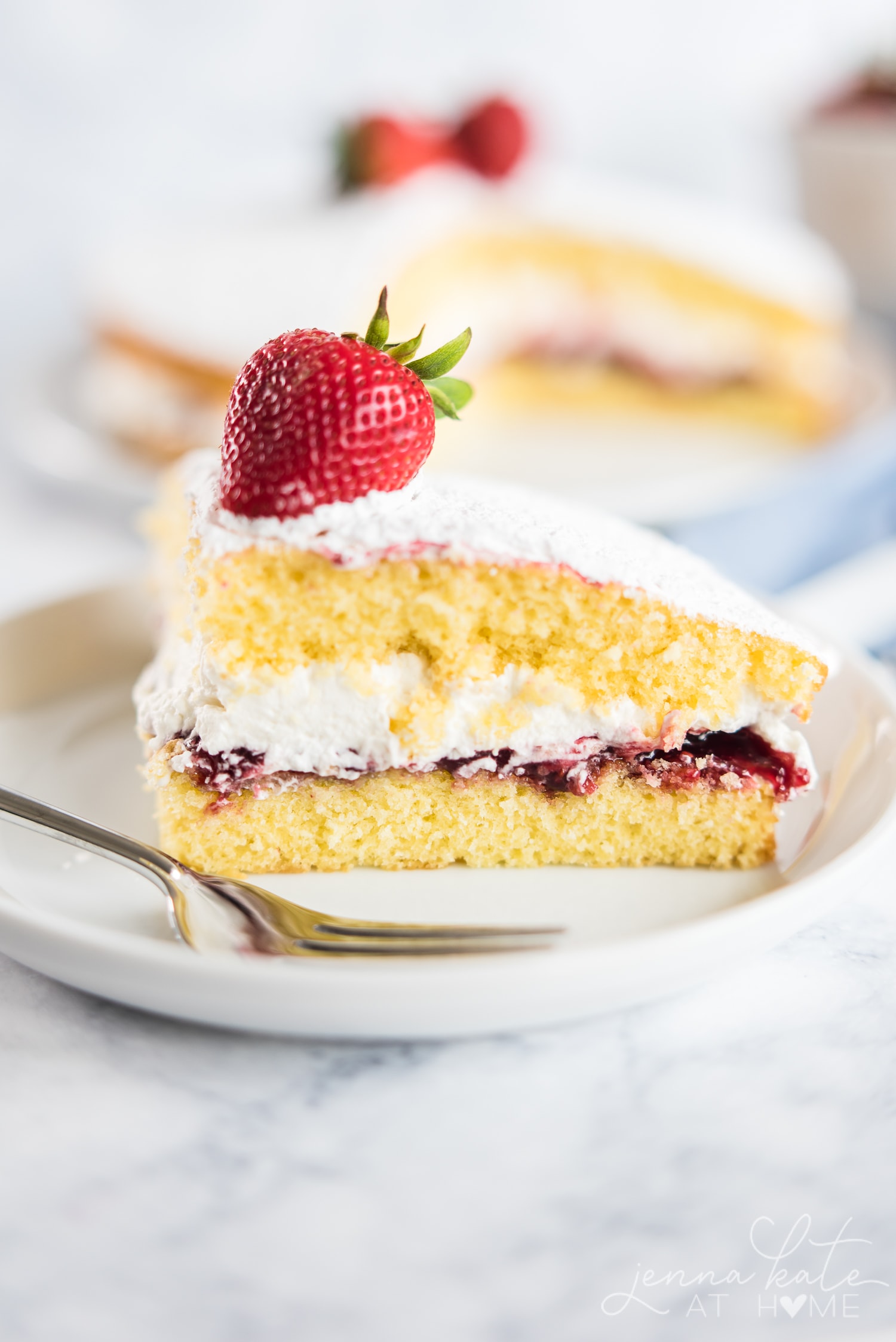 A slice of Victoria Sponge Cake is filled with a layer of raspberry filling and fresh whipped cream
